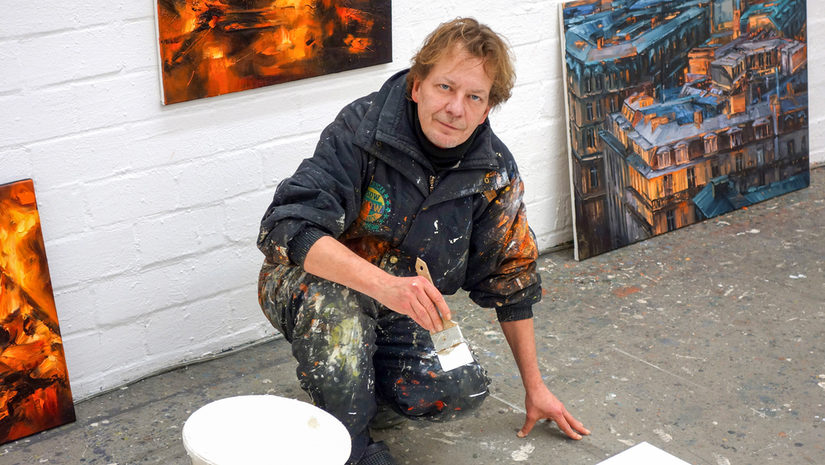 Das Bild zeigt den knieenden Thomas Prautsch, mit einem Pinsel in der Hand. Im Hintergrund stehen Malereien und auf dem Steinboden sind Farbkleckse