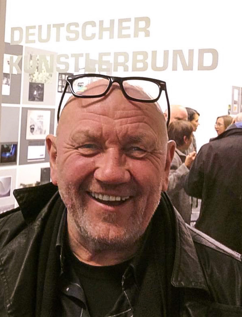 Ulrich Langenbach schaut lachend in die Kamera.Über ihm steht der Schriftzug Deutscher Künstlerbund. Im Hintergrund stehen mehrer Leute und unterhalten sich.