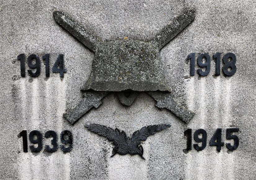 Detailaufnahme des Denkmals für gefallene Soldaten der beiden Weltkriege. Die Jahreszahlen der Kriege sind in schwarz auf grauem Stein aufgebracht. In der Mitte ein Helm und zwei überkreuzte Schwerter, darunter zwei Lorbeerzweige.