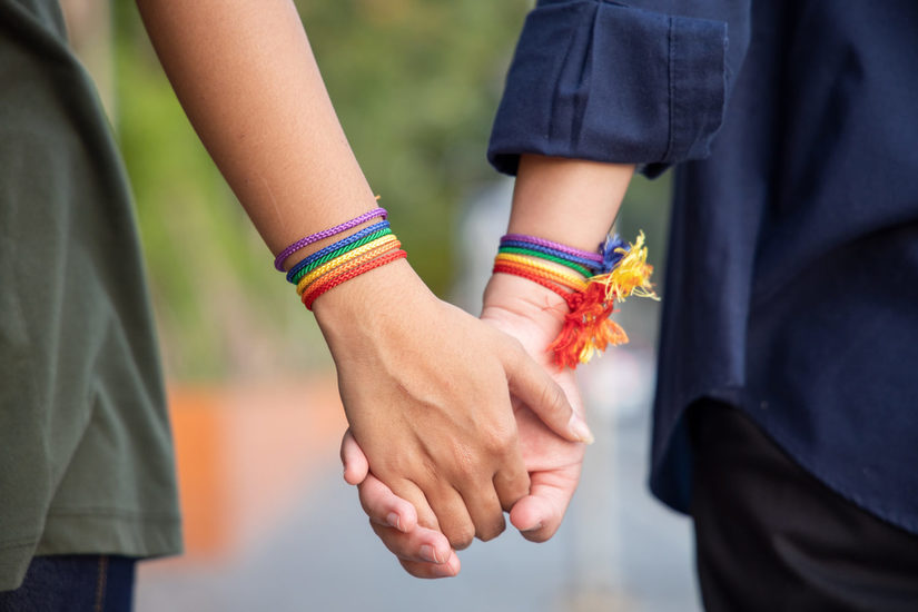 Zwei Personen mit regenbogenfarbenen Armbändern halten Hände.