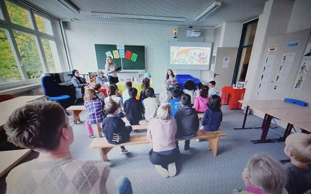 Ein Blick von hinten in den Musikraum, die Kinder sitzen auf Bänken und sind von hinten zu sehen. Vorn wird vorgelesen.