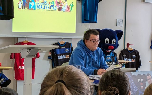 Herr Hornberger sitzt zusammen mit dem Maskottchen auf dem Stuhl und liest vor.