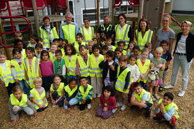 Auf dem Foto sind die Erstklässler mit ihren Lehrerinnen, der Schulleitung, der Polizistin und den Eltern, die den Walking Bus begleiten, zu sehen.