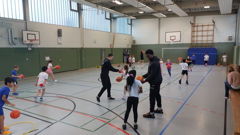 Kinder dribbeln in der Halle, die Basketballtrainer helfen ihnen dabei.