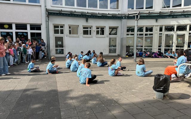Die Schülerinnen und Schüler sitzen in blauen Gemeinschaftsshirts auf dem Boden vor dem Hauptstandort und warten auf den Beginn des Tanzes.