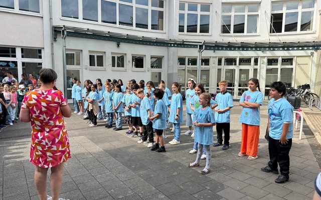 Frau Schäfers hält eine Rede. Sie trägt ein rotes Kleid. Die Schüler stehen in Reihen vor ihr und hören zu.