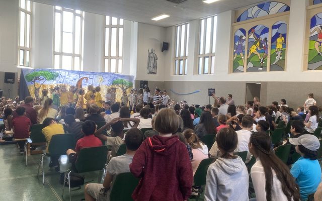 Die volle Aula ist zu sehen, im Hintergrund das Bühnenbild.