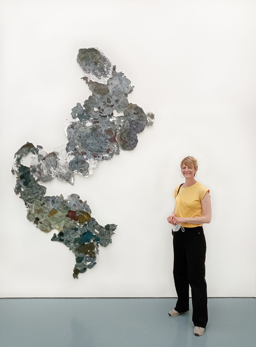 Verena Freyschmidt steht rechts von einer abstrakten Malerei. Die Malerei ist dreidimensional und grünlich,grau gefleckt.
