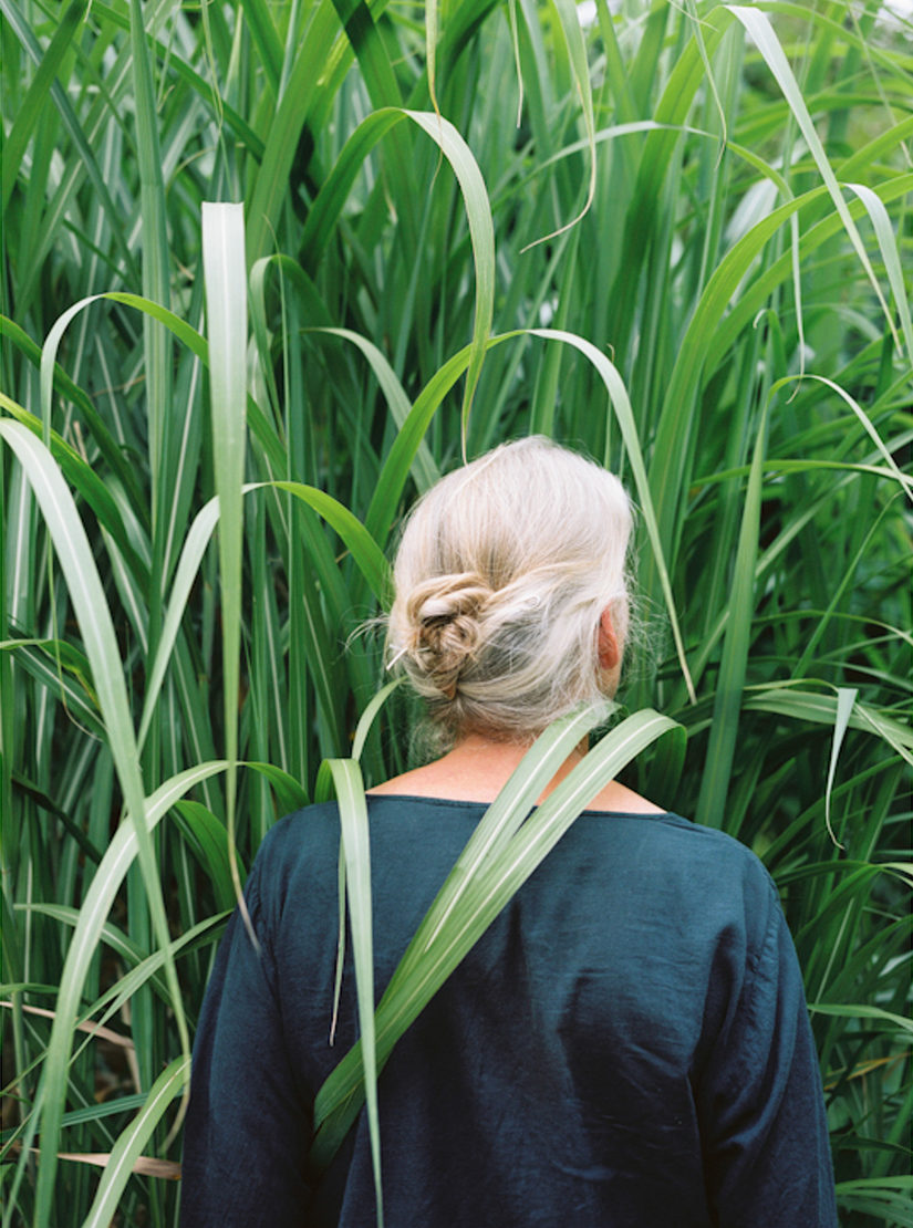 Eine blonde Frau steht mit dem Rücken zur Kamera in einem Feld. Das Gras ist höher als sie selbst.