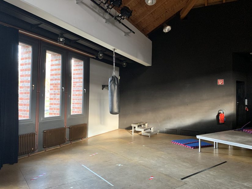Links auf dem Foto ist eine Fensterfront, daneben hängt ein schwarzer Boxsack sowie eine Klimmzugstange. Die Bühne des Saals ist rechts zu sehen.
