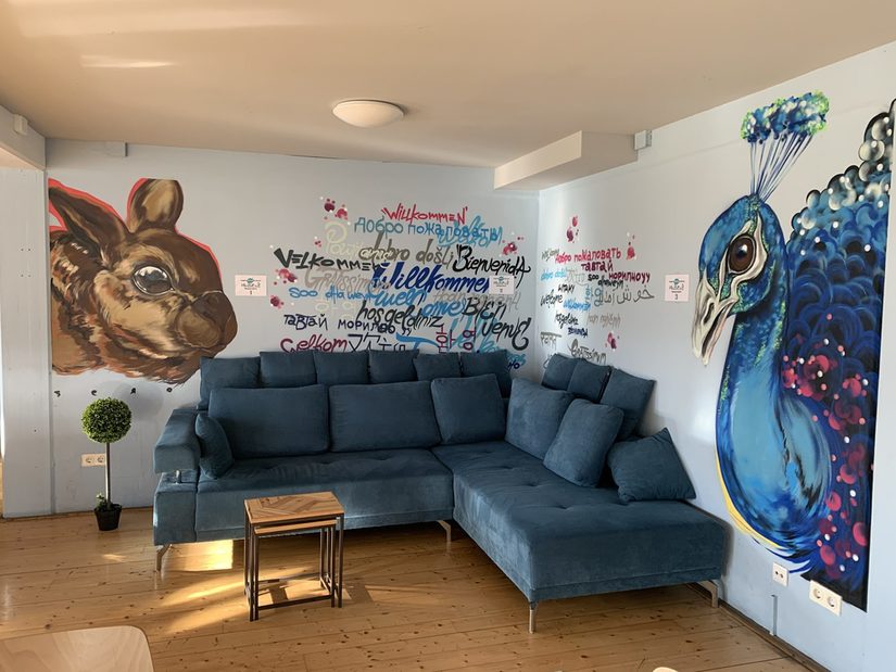 Das Foto zeigt ein petrolfarbenes Ecksofa. Die Wand hinter dem Sofa ist in hellblau gestrichen. Darauf steht mit Graffiti "Herzlich Willkommen" in unterschiedlichen Sprachen. Links an der Wand ist ein Hase gesprüht, rechts ein Pfau.