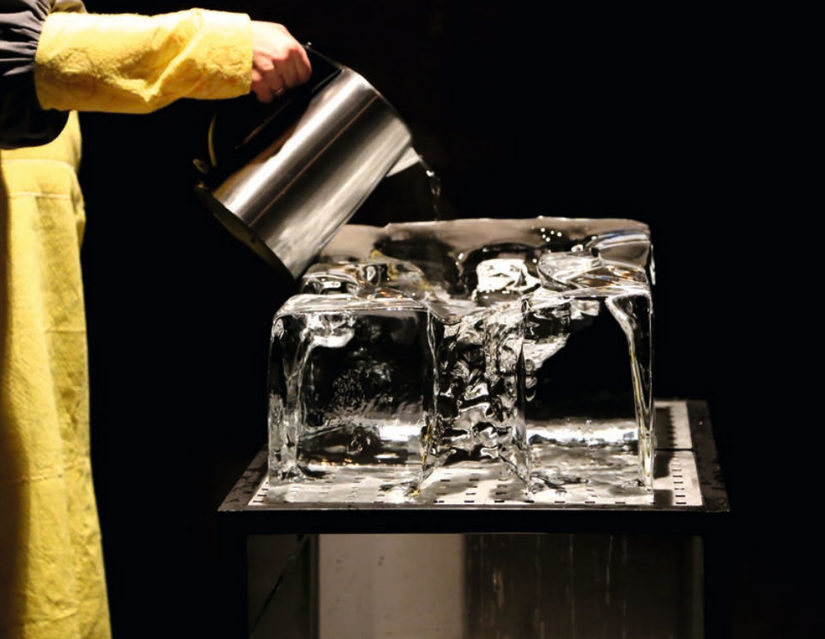 Eine Person mit einer gelben Schürze gießt Wasser aus einem silbernen Krug auf einen großen Eisblock