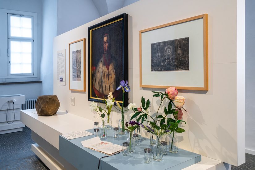 Foto aus der Ausstellung mit einem weißen Display, der Gemälde und einzelne Blumen in Gläsern zeigt.