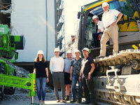 Auf dem Foto sind mehrere Projektverantwortliche zu sehen, die in, auf und um einen Bagger vor der Baustelle am Abdinghof stehen.