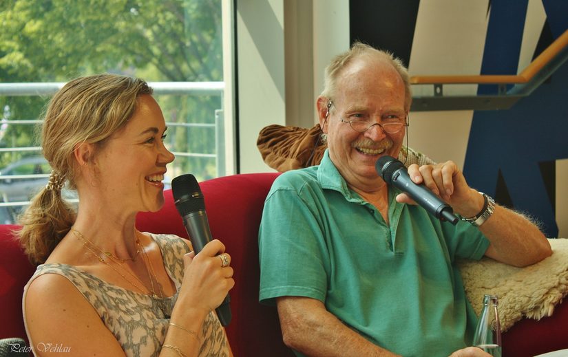 Modoratorin Julia Ures und Dave Lubek sitzen auf einem roten Sofa erzälen und lachen.