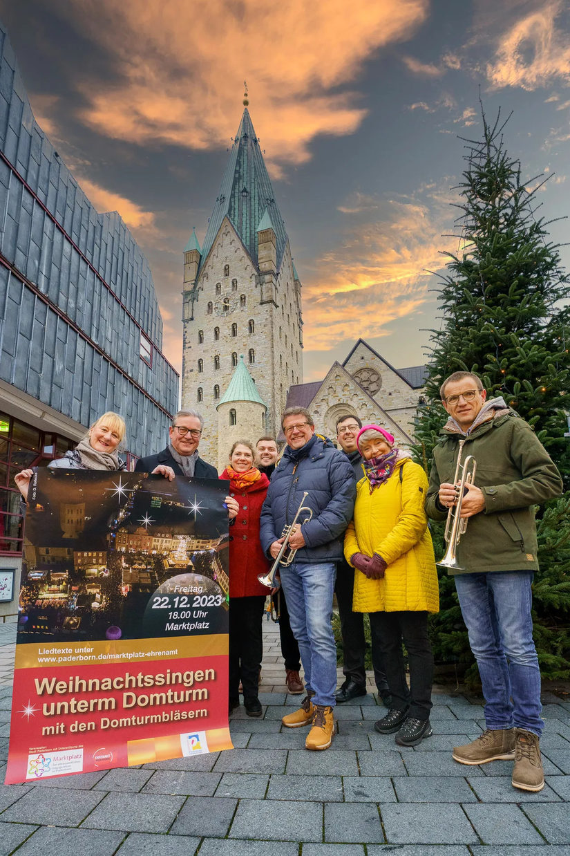 Verschiedene Personen positionieren sich vor dem Dom mit Blasinstrumenten und einem Plakat des Weihnachtssingen in der Hand