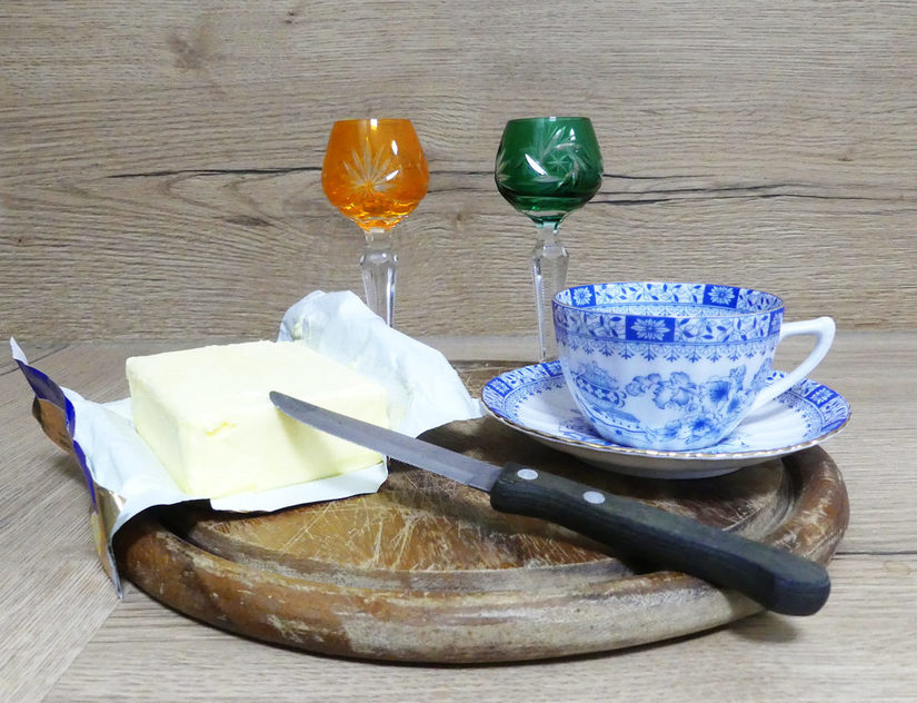 Holzbrett mit einem Stück Butter, Messer, zwei Gläsern und einer Kaffeetasse mit Untertasse