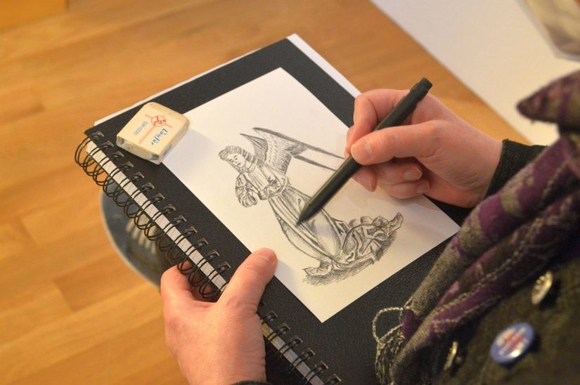 Ein Mensch hält einen Zeichenblock in der Hand und zeichnet mit Bleistift einen Engel