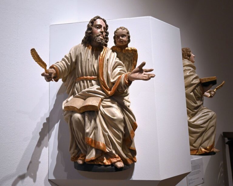 Zwei Figur aus dem Diözesanmuseum Paderborn werden gezeigt. Die Linke hält eine Feder und trägt beige-goldene Kleidung die rechte Person ist ein Kind